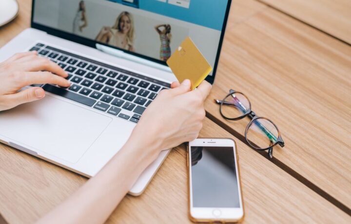 A person makes an online purchase using a credit card on a laptop, with a smartphone and glasses nearby.