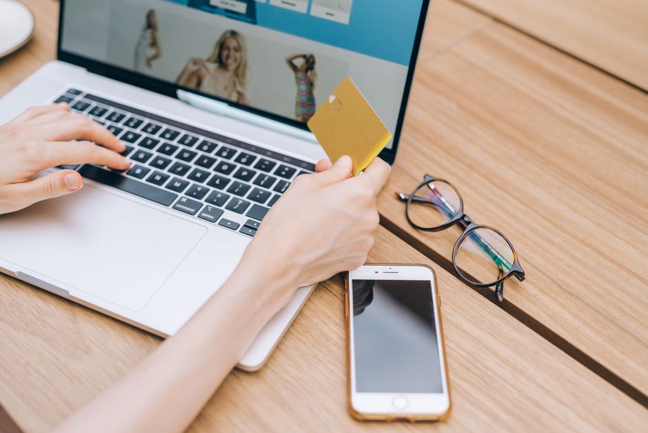 A person makes an online purchase using a credit card on a laptop, with a smartphone and glasses nearby.