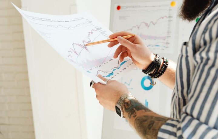 A man analyzing stock market charts with a pen, holding a paper report indoors.