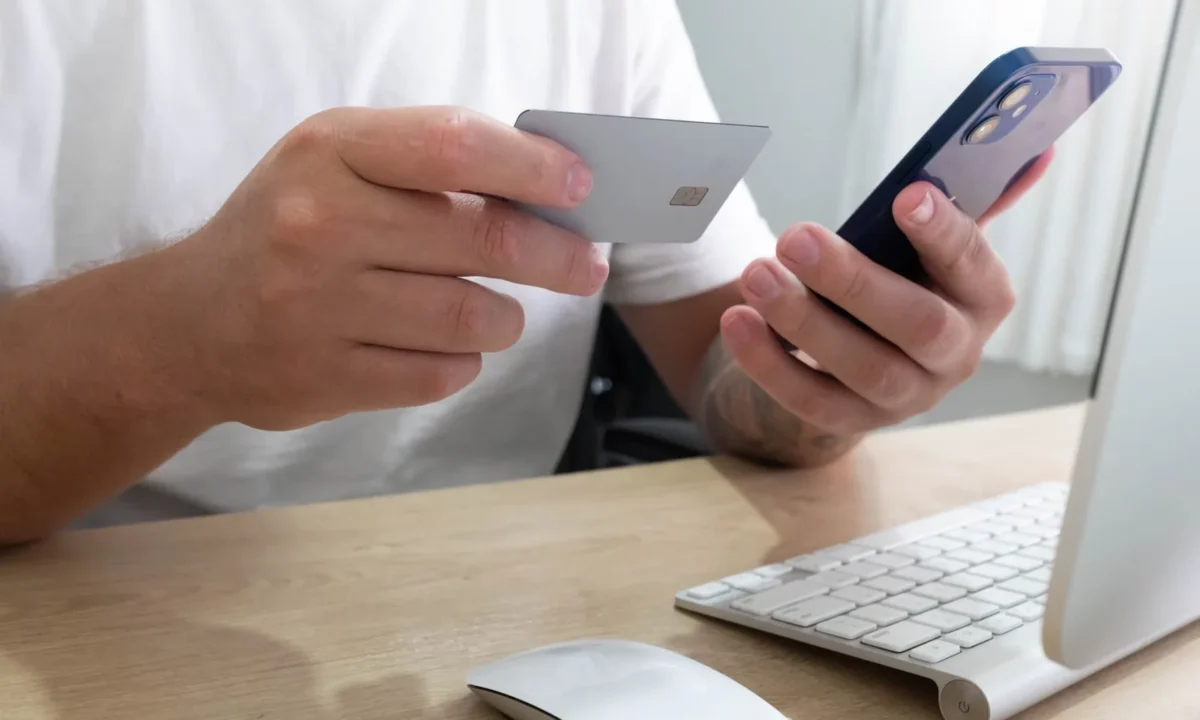 Pessoa segurando um cartão de crédito e um smartphone realizando uma transação online em uma mesa