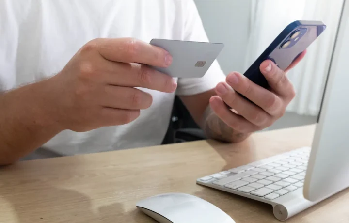Pessoa segurando um cartão de crédito e um smartphone realizando uma transação online em uma mesa